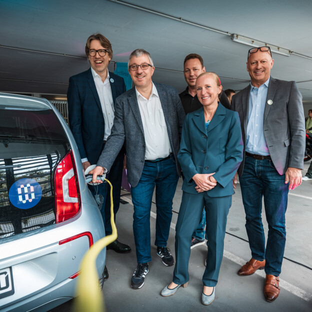 Gruppenbild mit 4 Männern und 1 Frau. Ein Mann hält den Ladestecker ans Auto. Auf dem Auto ist das bunte Münchner-Wohnen-Logo zu sehen.