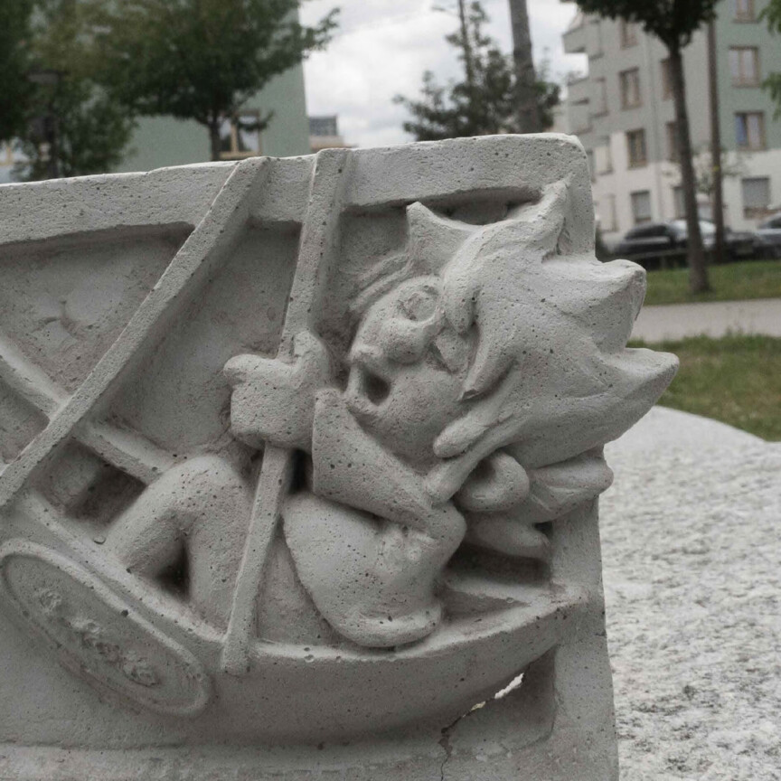 Skulptur aus Beton, die den Kobold Pumuckl in seiner Schiffsschaukel schaukelnd zeigt.