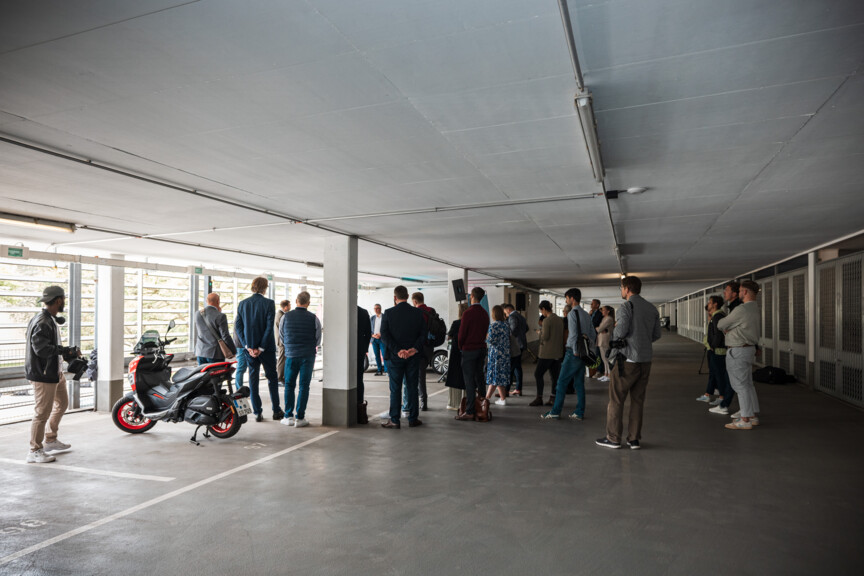 Eine Gruppe von Menschen stehen in einer Garage im Kreis und hören einem Redner zu. Links sind Fenster zu sehen und Mofa.