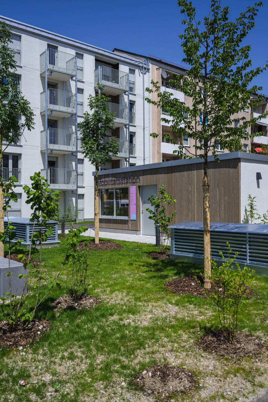 Ansicht des Wohnhauses Ittlinger Straße. Im Vordergrund die grünen Freiflächen mit der Mobilitätsstation. Im Hintergrund das Wohnhaus mit Balkonen.