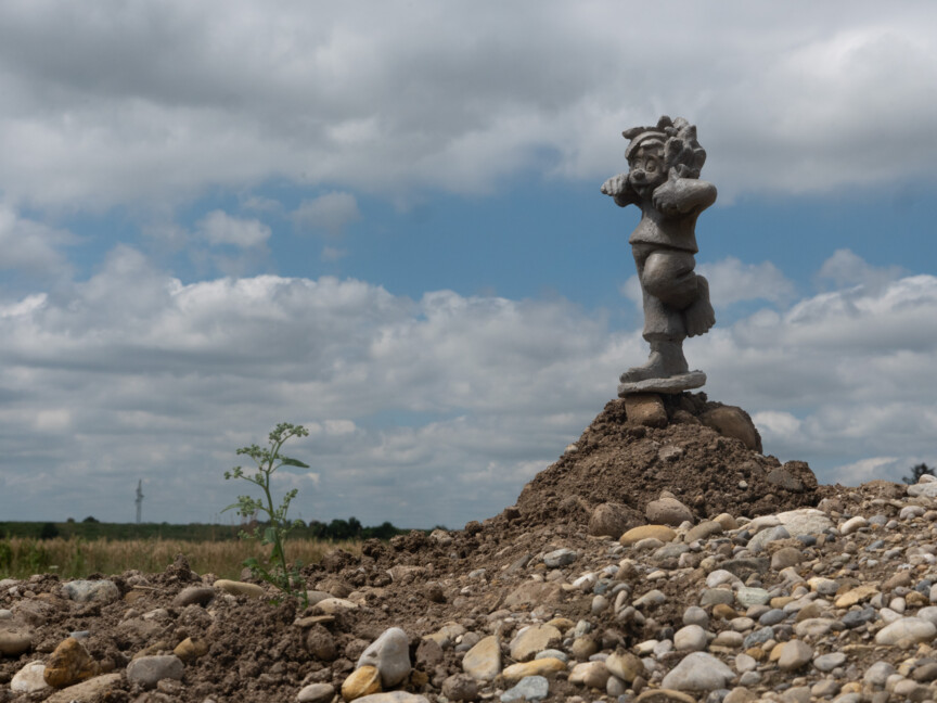 Foto einer Betonfigur von Pumuckel. Der Kobold steht auf einem Bein auf einen Berg aus Sand und Steinen. Im Hintergund ein Feld.