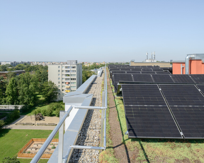 Blick von oben auf das Dach einer Wohnanlage, auf dem eine Photovoltaikanlage installiert ist. In der Ferne ist die Skyline von München zu sehen. Links vom Gebäude eine Grünanlage und ein weiterer Wohnkomplex.