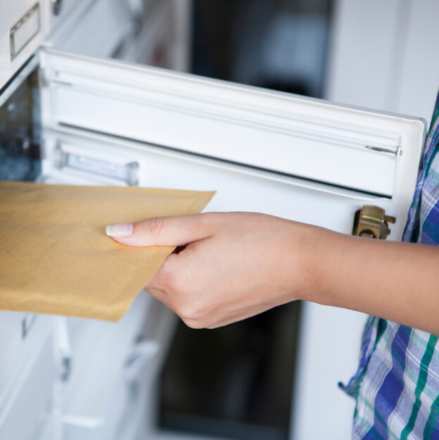 Man sieht eine Hand, die einen Din A4 großen, braunen Briefumschlag aus dem Briefkasten nimmt.