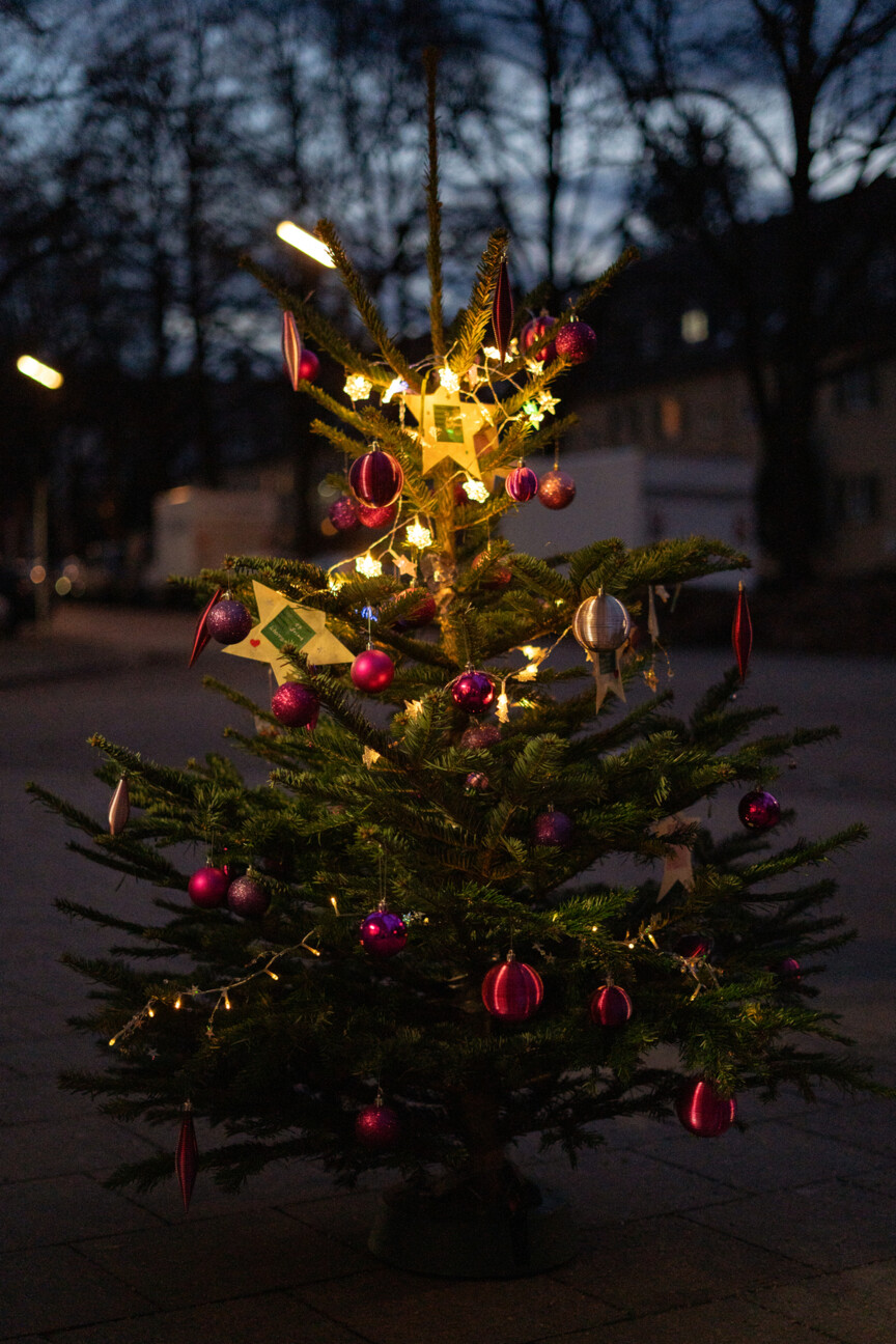 Im Fokus steht ein geschmückter Weihnachtsbaum. Im Hintergrund sieht man einen Wohnblock in der Abenddämmerung.